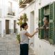 Young woman giving water to an elderly neighbour at a shuttered window in a white Andalusian street on a hot summer morning